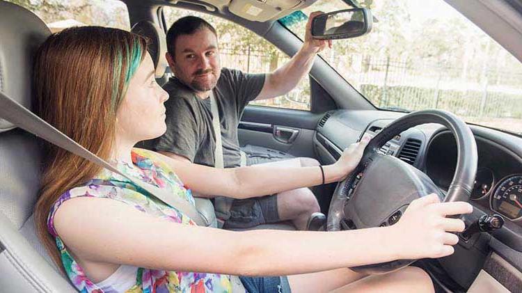 Teen girl in the driver's seat of a car, buckled up with hands on the wheel. Her father is in the passenger seat adjusting the rearview mirror. 